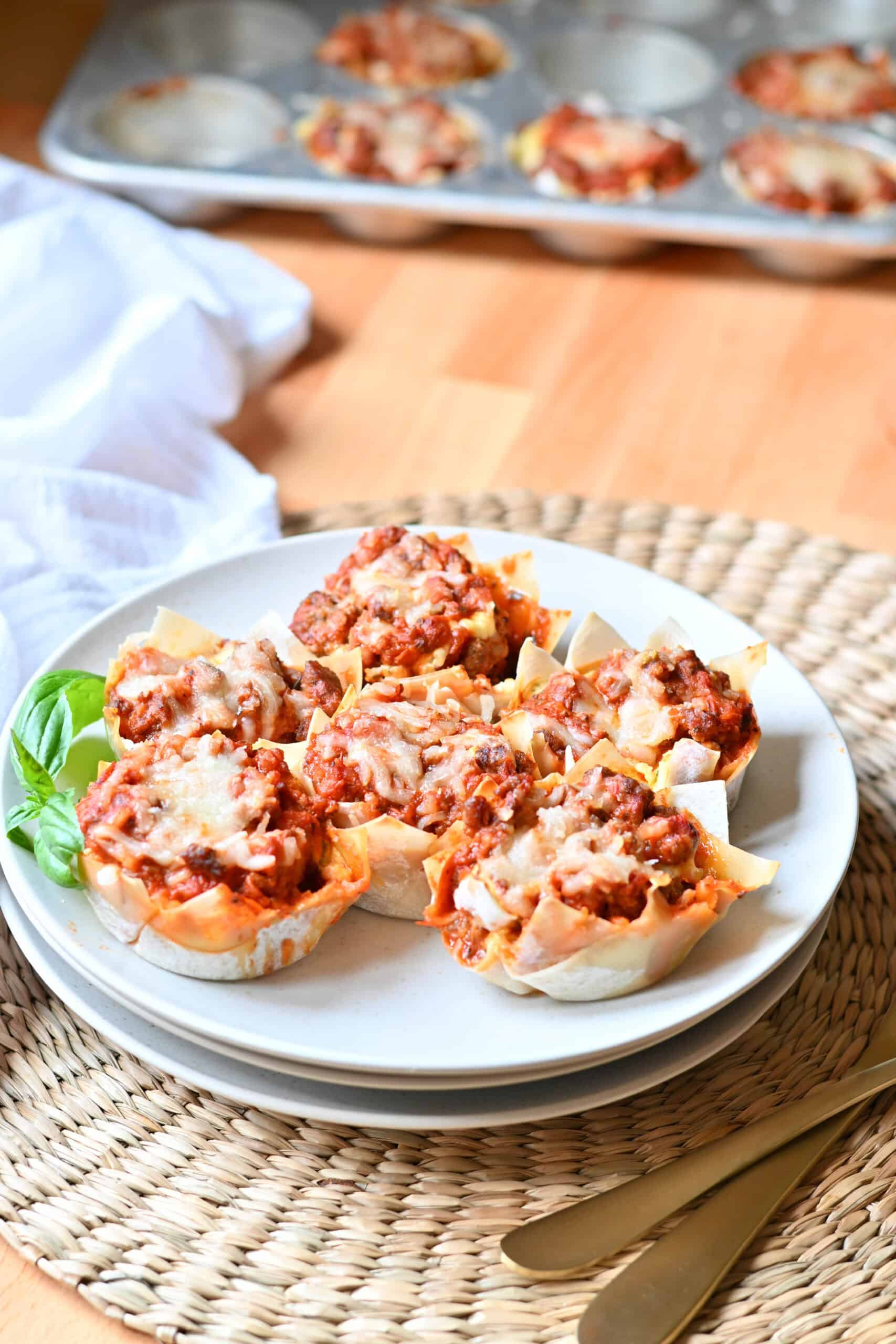 Plate of six lasagna cups with a muffin tin of more lasagna cups behind it.